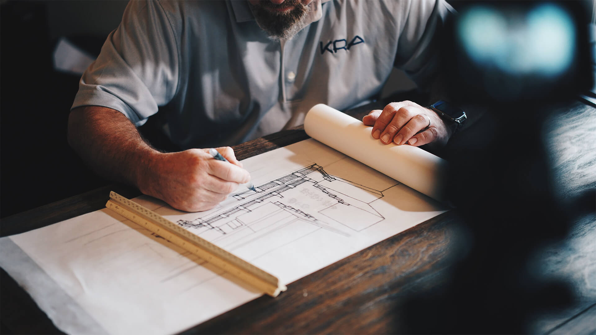 Architect drawing a detailed building section sketch with a ruler and pencil on rolled drafting paper at a wooden desk