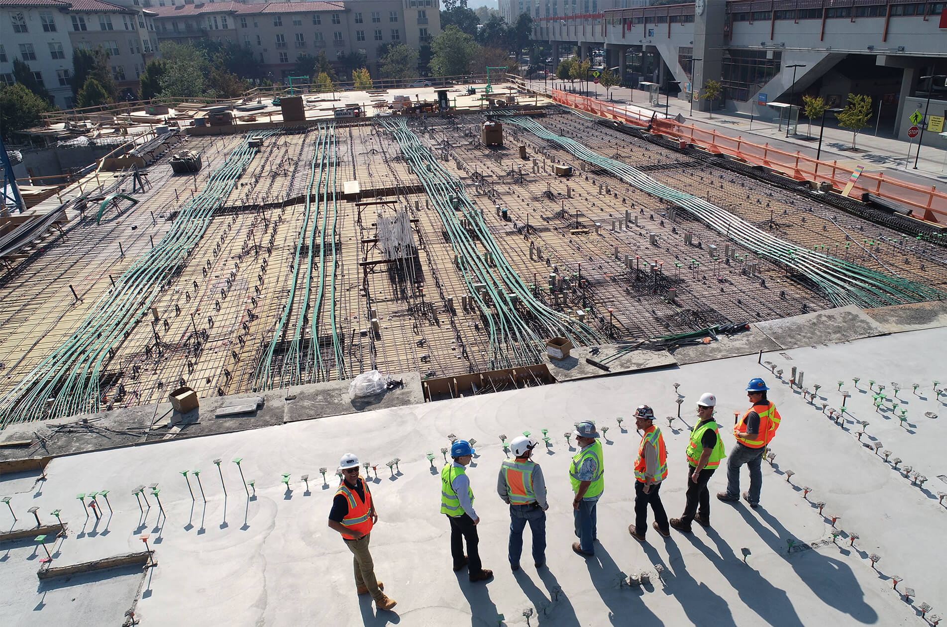 Construction workers in safety vests and hard hats standing on a concrete slab overlooking a large rebar foundation site with post-tension cables