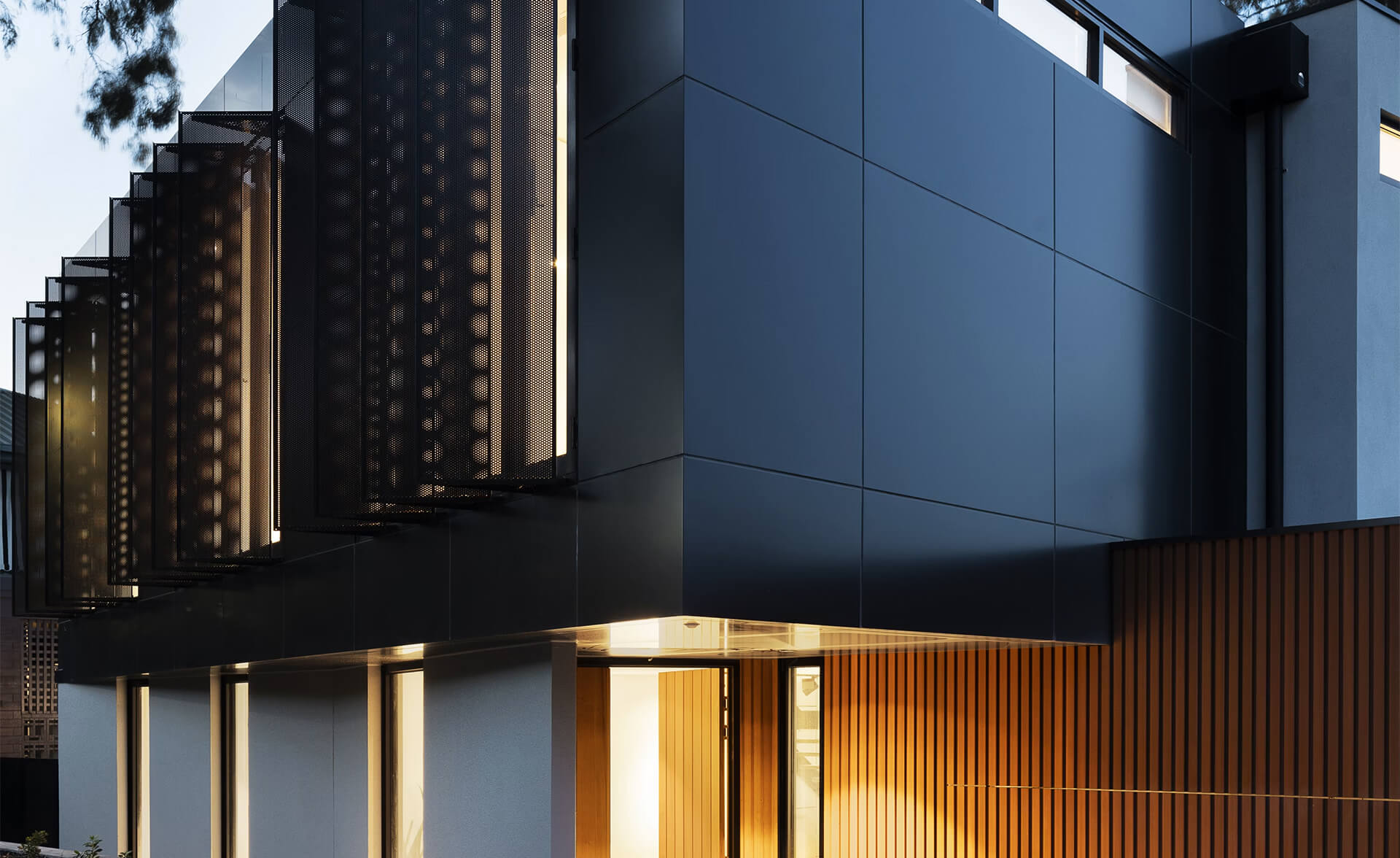 Close-up exterior of a contemporary home at dusk with navy cantilevered upper floor, perforated metal shutters, and glowing timber entrance below