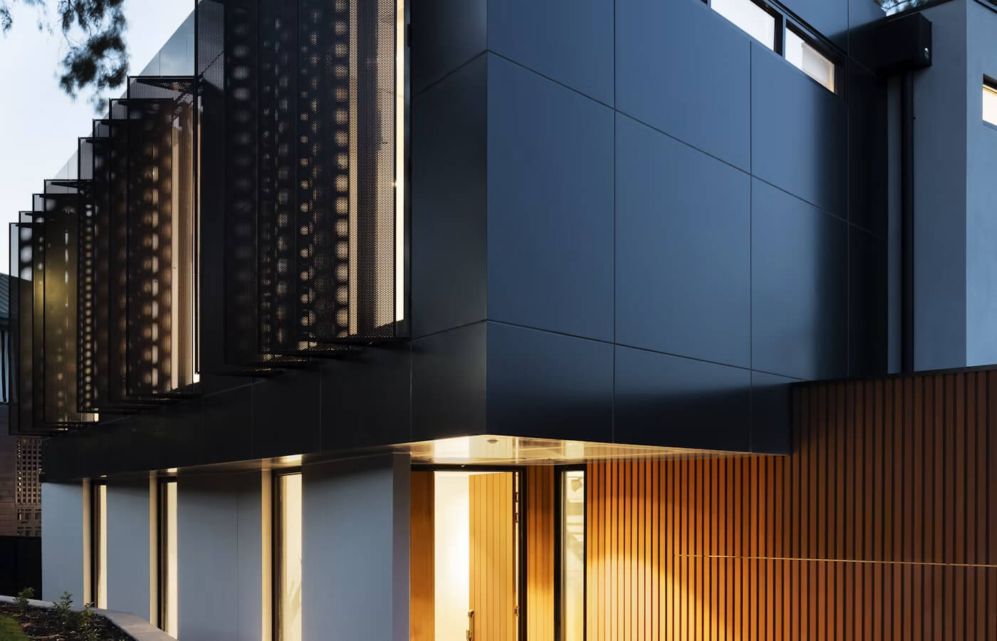 Modern house exterior at dusk with dark blue cladding, perforated metal shutter panels, and a warmly lit entrance with wood slat wall