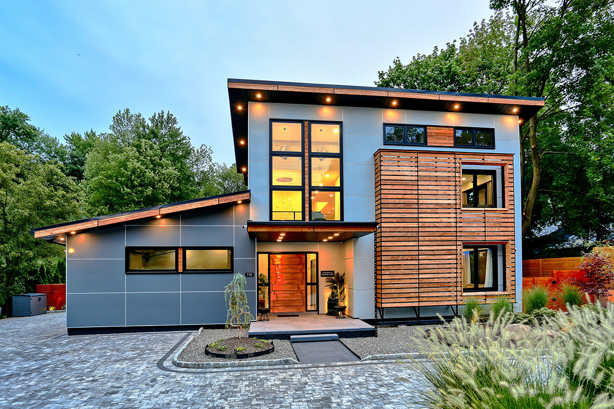 Contemporary two-story home at twilight with warm interior lighting, wood slat cladding, gray panels, and a landscaped stone driveway