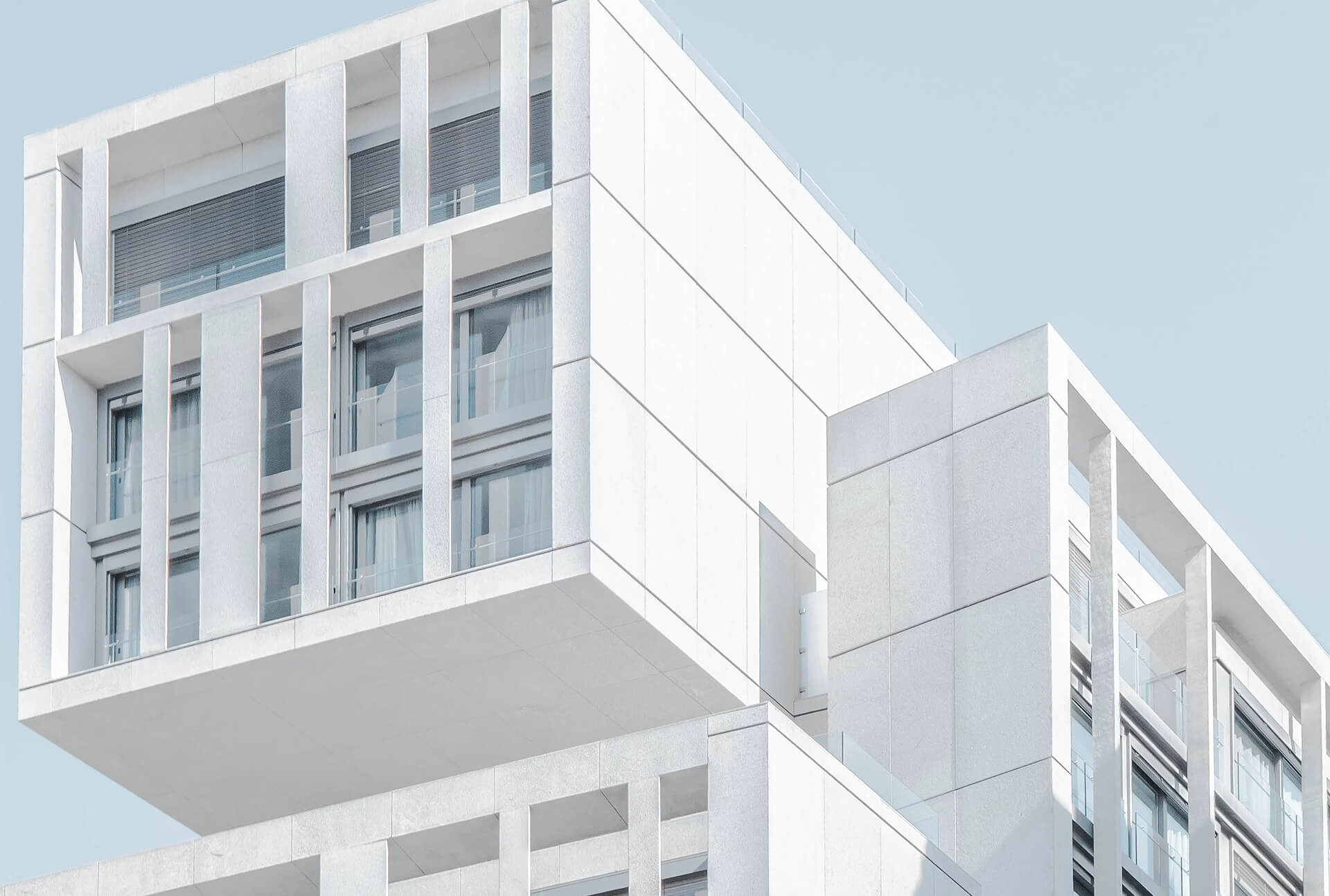 Low-angle view of a modern white building with cantilevered balconies, geometric cladding, and large windows against a clear sky