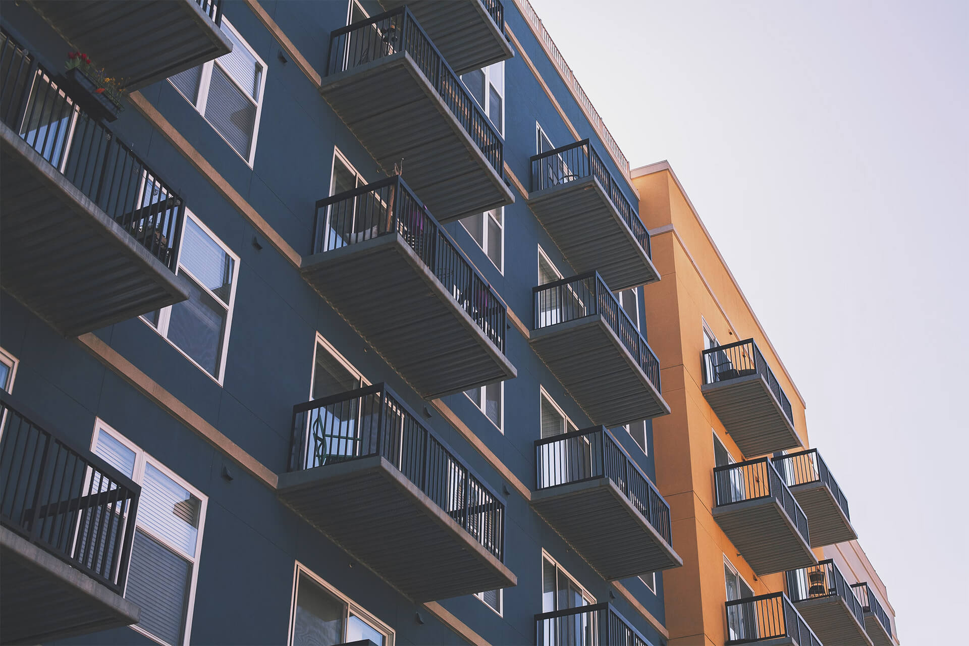 Angled exterior shot of a multi-story apartment complex with tiered metal balconies along blue and orange facades