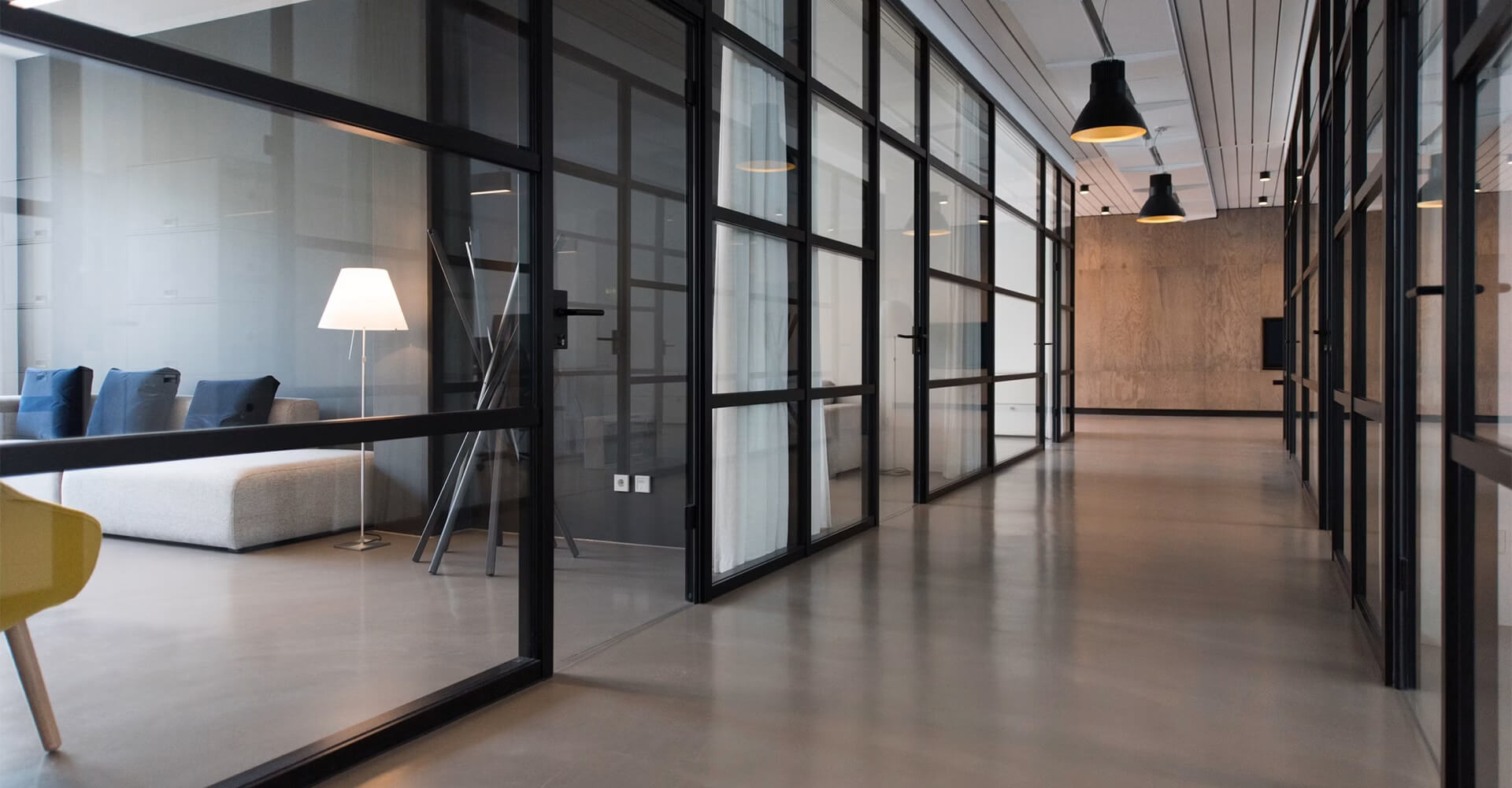 Modern office corridor with black-framed glass partition walls, pendant lighting, and a lounge area with sofas visible through the glass